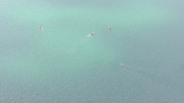 An aerial bird's-eye view of five kayakers paddling across the vast calm turquoise water of the Quilotoa crater lake in the Ecuadorian Andes, gentle ripples spreading on the clear surface.