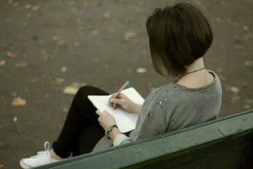 Brunette girl siting on the park bench and writing notes into her notepad