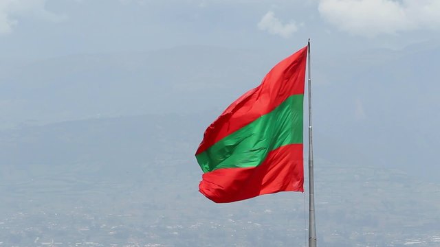 A red and green provincial flag of Tungurahua waves in the wind atop a pole, high above the city of Ambato and the distant Andes mountains under a cloudy sky, Ecuador.