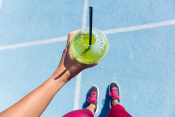 Runner drinking a healthy spinach green smoothie on outdoor running track getting ready for run. Closeup of hand holding juice drink on blue lane, social media health and fitness concept.