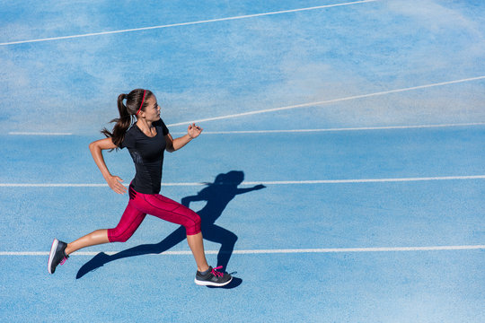 Athlete runner running on athletic track training her cardio. Jogger woman jogging at fast pace for competition race on blue lane at summer outdoor stadium wearing red capri tights and sports shoes.