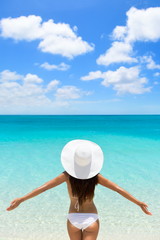 Happy beach vacation carefree woman on luxury tropical getaway destination wearing a white sun hat from behind with arms raised in freedom.  Person from behind with slim body. Happiness and success.