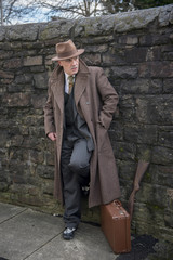 Vertical portrait image of a mature man dressed as a 1940s gangster character, with a suitcase and a shot gun.