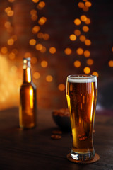 Glass of light beer with bottle and snacks on wooden table over glitter background