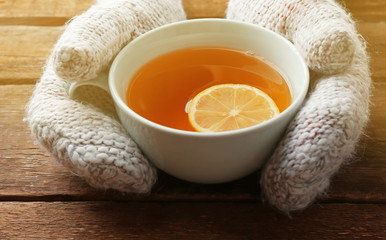Female hands in mittens holding cup of tea on wooden background