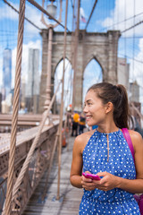 Fototapeta premium Happy Asian woman using phone texting walking on famous attraction landmark Brooklyn bridge, New York city for social media. Tourist sightseeing on summer travel in Manhattan, USA.