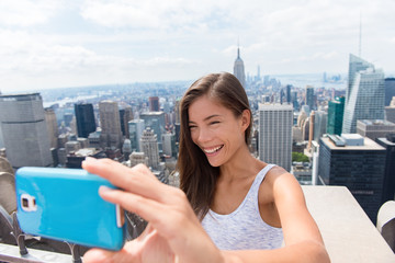 Tourist woman taking mobile phone selfie picture at New York skyline. Asian girl holding blue smartphone for self-portrait photo with view of Manhattan skyscrapers during summer travel vacation.