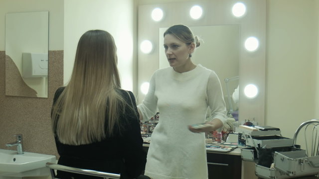 Professional makeup artist talking and applying makeup powder on a cheek on face of client female, opposite mirror with light