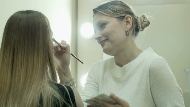 Professional makeup artist talking and applying makeup powder on a cheek on face of client female, opposite mirror with light