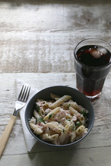 Pasta penne is cooked al dente, then mixed with sliced ham, feta cheese and basil. Dressing is made with olive oil, bit of vinegar, salt and pepper. Served with glass of soda on white rustic table. 
