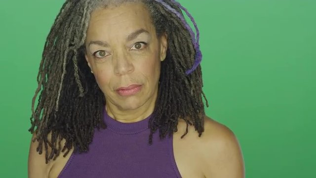 Older African American woman with dreadlocks looks upset and then laughs, on a green screen studio background