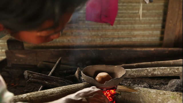 A person tends a small clay pot over glowing embers in a rustic open-air kitchen, using a stick to adjust the fire, traditional cooking with natural materials in the Ecuadorian Amazon.