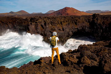 Male tourist photographing Los Hervideros rocky coast with wavy ocean and volcanos on the background on Lanzarote island in Spain