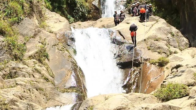 A group of adventurers in safety gear rappel down a rocky waterfall cascade in a lush tropical canyon, one descending as others wait on the ledge above, near Banos, Ecuador.