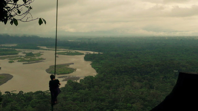 A person swings on a rope from a high vantage point over a vast tropical jungle and winding river in the Ecuadorian Amazon, silhouetted against an overcast sky in a remote lush environment.