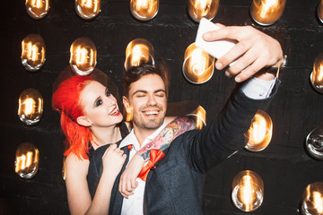 Happy couple in nightclub do selfie on mobile phone. Young redheaded girl with big tattoo hugs young laughing men. Party selfie of boyfriend and girlfriend on black background.