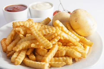 Portion of French fries (Crinkle-cut) deep fried, served on a white plate next to white bowls with mayonnaise and ketchup and fresh potato.