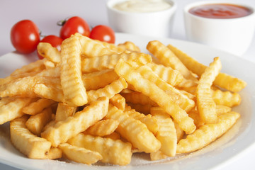 Portion of French fries (Crinkle-cut) deep fried, served on a white plate next to white bowls with mayonnaise and ketchup.