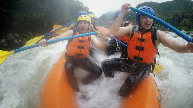 A group of adventurers in an orange raft navigate fast-moving rapids near Banos, Ecuador, paddling vigorously and reacting to the churning water in a close dynamic onboard shot.