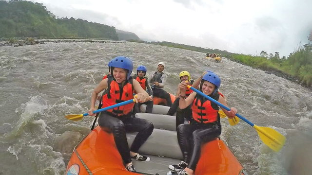 A group of adventurers in an orange raft navigate turbulent river rapids near Banos, Ecuador, paddling with yellow oars in helmets and life vests, a front-facing shot of splashing water.