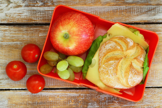 Lunch box containing cheese sandwich, red apple, grapes and cherry tomatoes on wooden surface
