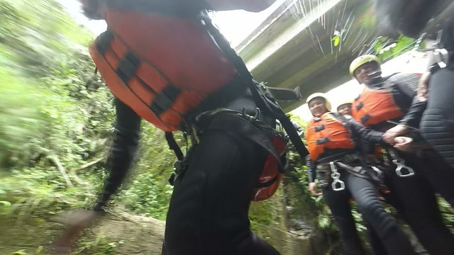 A group of adventurers in safety gear prepare and jump into a rocky waterfall pool in a lush jungle canyon near Banos, Ecuador, the jump from low angle to a wide shot of the cascade.