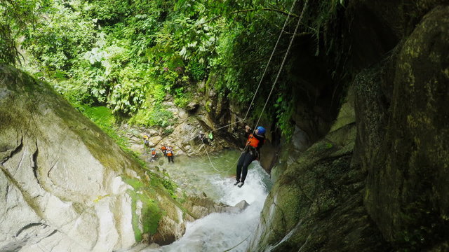A group of adventurers navigate a lush moss-covered canyon with a rushing river below, one descending by rope, canyoning in dense green jungle near Banos, Ecuador.