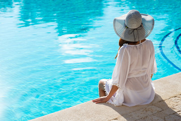 Fototapeta na wymiar Back view of fashion woman on summer vacation relaxing at luxury resort spa poolside. Young fashionable lady wearing sun hat and white kaftan.