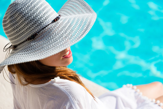 Fashion Pretty woman on summer vacation relaxing at luxury hotel resort spa poolside. Young  fashionable lady wearing sun hat and white kaftan.