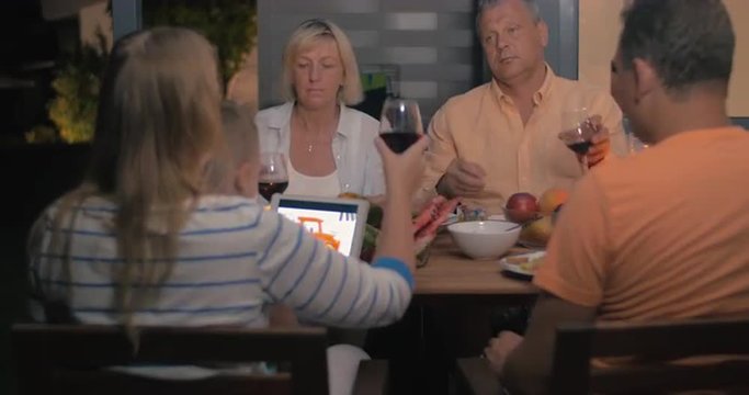Outdoor family supper in late evening. Parents and grandparents toasting with wine and having meal while child watching cartoons on tablet computer