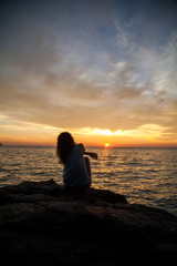 a young girl sits with his back turned on the rock during sunset by the sea