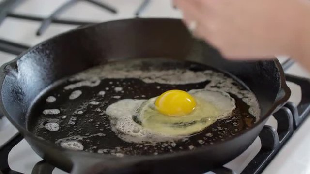 Cook's hands crack an egg and drop it into a cast iron pan, where it starts frying on a gas stove.  Hand held camera, recorded at 4K UHD.