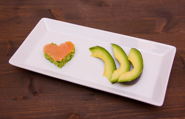 Slices of avocado and avocado and smoked salmon in the shape of heart on tray on wood