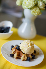 Delicious dessert at home. A big Belgian waffle with a scoop of vanilla ice-cream and organic blueberries on a white plate on a yellow tablecloth.
