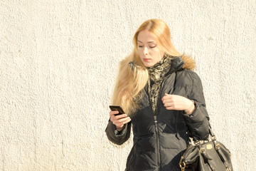 Close up portrait of a young fashionable woman in a black parka on the street near the wall holding a smartphone © elen31