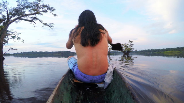 Capture the breathtaking beauty of a stunning sunset as indigenous men gracefully navigate a wooden canoe,creating a mesmerizing scene from a captivating backside camera angle.