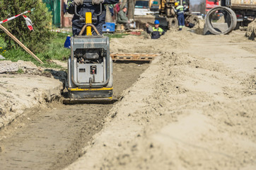 A worker with a vibrating plate in a ditch with a workers and machinery in background
