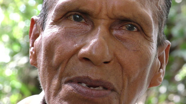 A close-up of an elderly Siona indigenous man with weathered skin and expressive eyes speaking outdoors in the Cuyabeno Amazon, Ecuador, against a natural green background.