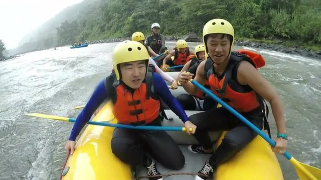 A group of adventurers in yellow helmets and orange life vests navigate a turbulent river in a yellow raft near Banos, Ecuador, paddling through rapids in a front-facing low-angle shot.