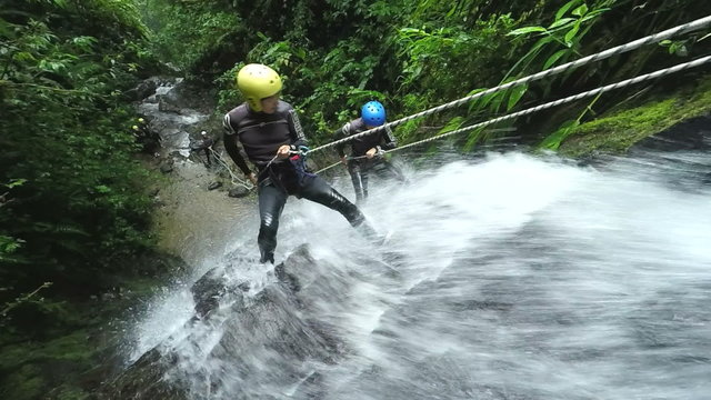 A group of adventurers in helmets and wetsuits descend a rocky rushing waterfall in lush tropical jungle near Banos, Ecuador, using safety ropes through the dense green forest.