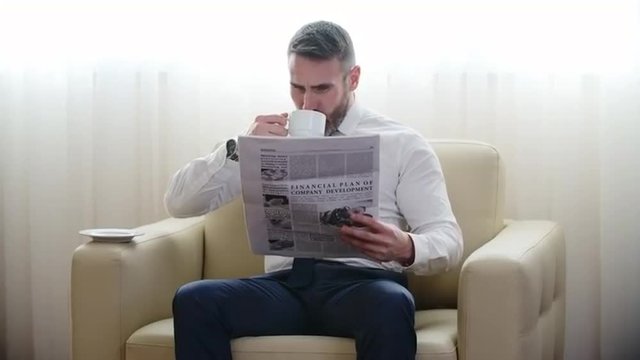 Mature businessman sitting in armchair, drinking coffee and reading newspaper