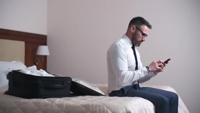 Businessman sitting on bed in hotel room and using his mobile phone