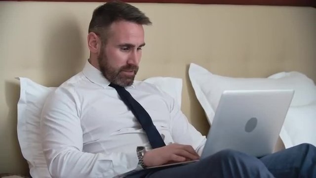 Businessman lying on bed with laptop, typing and smiling