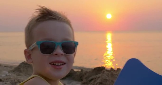 Close-up shot of a blond little boy in colorful sunglasses on background of quiet sea and golden sunset. Summer holidays