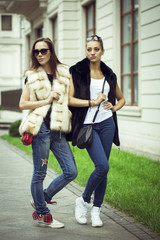 Fototapeta premium Fashion shot of two elegant beautiful girls in the sunset wearing sunglasses, fur vests . Two young women outdoor on the street. Shopping inspiration