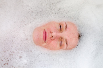 Face of a young beautiful girl in a white bath among soap bubbles from the foam bath gel, naked with wet hair, enjoys the spa treatments for beauty and health
