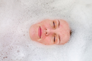 Fototapeta premium Face of a young beautiful girl in a white bath among soap bubbles from the foam bath gel, naked with wet hair, enjoys the spa treatments for beauty and health