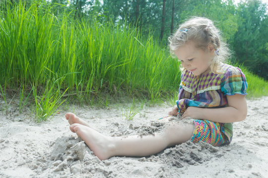 Little girl playing on beach dune and burying herself in white sand at summer pinewood background