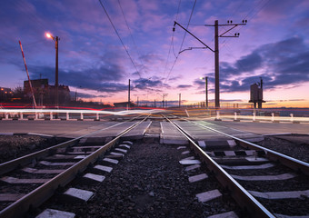 Fototapeta na wymiar Rail crossing with blurred car lights on the background of colorful cloudy sky at beautiful sunset. Railway landscape