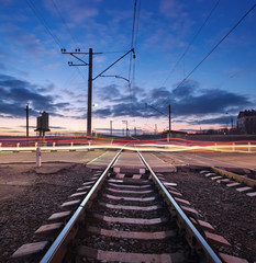 Fototapeta na wymiar Rail crossing with blurred car lights on the background of colorful cloudy sky at beautiful sunset. Railway landscape